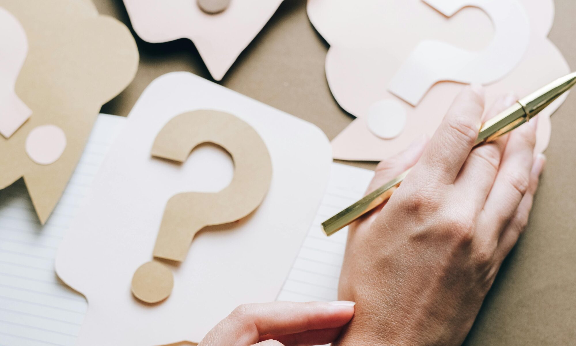 Photo of paper question marks with hands and a pen suggesting curiosity and inquiry.